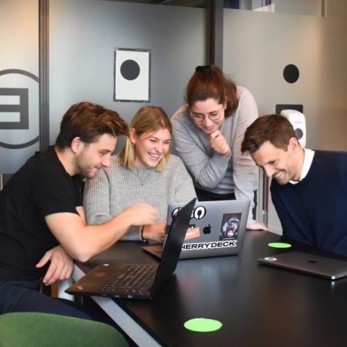 Personer som sitterog stårforann en PC